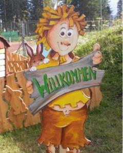 Eine Zeichentrickfigur mit einem Schild mit der Aufschrift "Willkommen" und einem Kaninchen, das hinter der Figur hervorlugt, steht im Freien auf einer Wiese mit einem Zaun und Bäumen im Hintergrund.