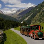 Ein grün-roter Touristenzug fährt auf einem gepflasterten Weg durch ein grünes Tal, umgeben von Bergen und Wäldern unter blauem Himmel.