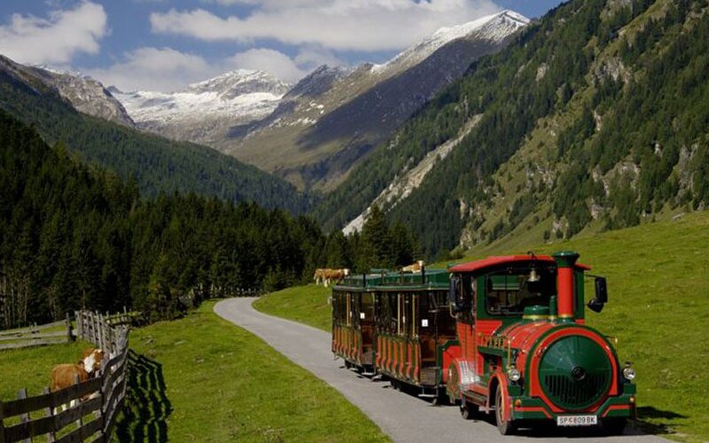 Ein grün-roter Touristenzug fährt auf einem gepflasterten Weg durch ein grünes Tal, umgeben von Bergen und Wäldern unter blauem Himmel.