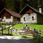 Zwei Steinhäuschen mit Holzdächern stehen an einem Holzzaun auf einer von Bäumen umgebenen Wiese. Eine Person steht in der Nähe des Zauns im Sonnenlicht.