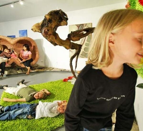 Kinder spielen auf Kunstrasen, während ein Mädchen an einer großen künstlichen Sonnenblume riecht; Erwachsene sitzen und unterhalten sich im Hintergrund einer Innenausstellung oder eines Kunstraums.