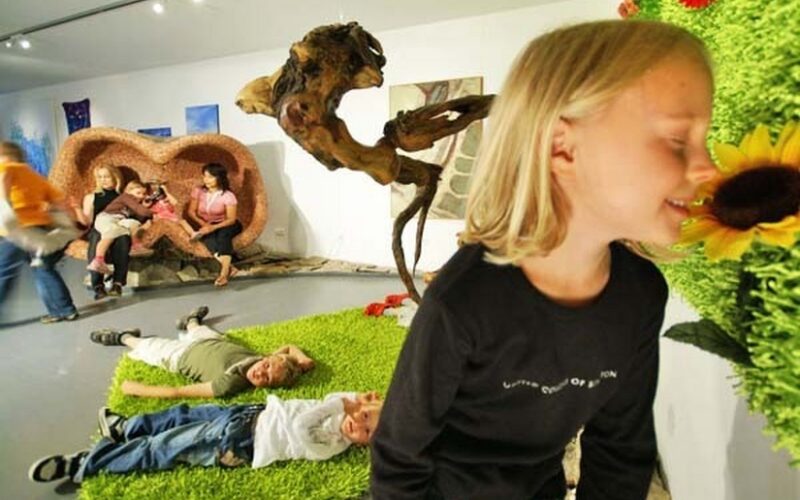 Kinder spielen auf Kunstrasen, während ein Mädchen an einer großen künstlichen Sonnenblume riecht; Erwachsene sitzen und unterhalten sich im Hintergrund einer Innenausstellung oder eines Kunstraums.