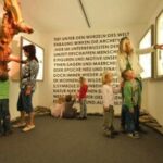 In einem hell erleuchteten Museumsraum mit Text an der Rückwand betrachten Kinder und Erwachsene Schautafeln und eine Baumausstellung.