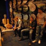 Drei junge Männer sitzen und stehen in einem schwach beleuchteten Raum mit einer gemusterten Wand und abstrakten Kunstelementen im Hintergrund.