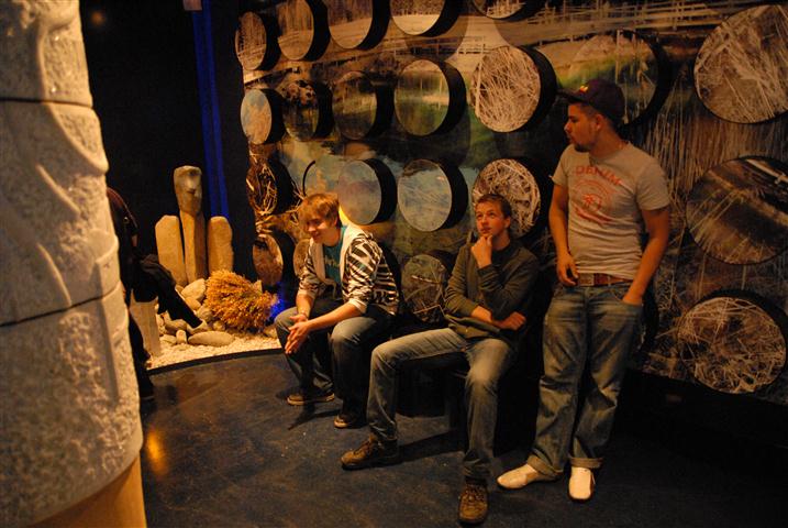 Drei junge Männer sitzen und stehen in einem schwach beleuchteten Raum mit einer gemusterten Wand und abstrakten Kunstelementen im Hintergrund.