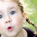 Kind mit hellbraunem Haar in Zöpfen und großen grünen Augen, die mit dem Mund ein "O" formen, im Freien vor einem verschwommenen grünen Hintergrund.