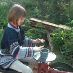 Ein Kind formt Ton auf einer Töpferscheibe im Freien, umgeben von Grünpflanzen und Holzstämmen.