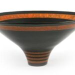 Eine breite, schwarze Keramikschale mit schmalem Sockel und orangefarbenen Zierbändern und -mustern am Rand und Boden.