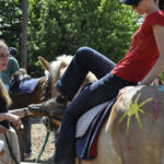 Eine Frau in einem roten Hemd besteigt ein bemaltes Pferd, während zwei andere Frauen in der Nähe im Freien stehen; eine hat einen Armgips, und im Hintergrund sind Bäume zu sehen.
