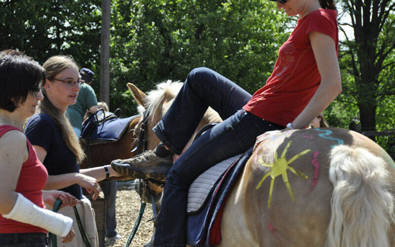 Eine Frau in einem roten Hemd besteigt ein bemaltes Pferd, während zwei andere Frauen in der Nähe im Freien stehen; eine hat einen Armgips, und im Hintergrund sind Bäume zu sehen.