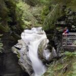 Eine Person steht auf einer hölzernen Plattform mit Blick auf eine enge Schlucht mit einem schnell fließenden Wasserfall inmitten eines dichten Waldes.