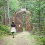Eine Person geht auf ein offenes Holztor in einem hohen Zaun zu, der von einem dichten grünen Wald umgeben ist.