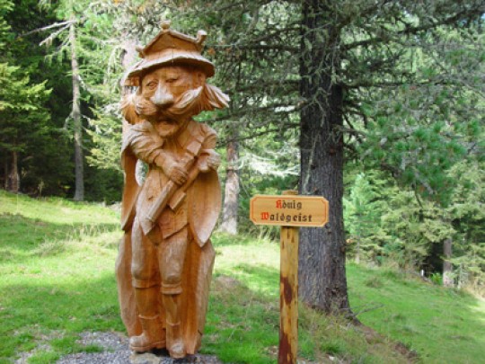 Holzstatue eines bärtigen Mannes mit Krone und Stab in der Hand, der in einem Wald neben einem Schild mit der Aufschrift "König Waldgeist" steht.
