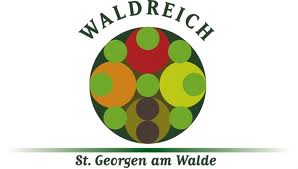 Logo für Waldreich St. Georgen am Walde mit einem kreisförmigen Design mit verschiedenen grünen, roten und orangefarbenen Kreisen und dem Namen ober- und unterhalb der Grafik.