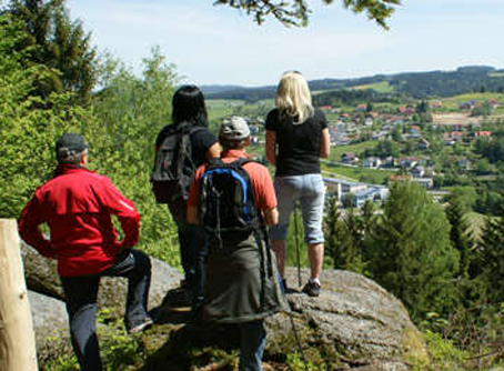 Vier Personen mit Rucksäcken stehen auf einem Felsen und blicken auf eine grüne Landschaft und eine kleine Stadt in der Ferne.