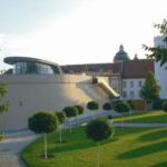 Modernes Gebäude mit geschwungener Architektur neben einem traditionellen Bauwerk, umgeben von einer Rasenfläche mit kleinen, runden Bäumen und einem gepflasterten Weg.