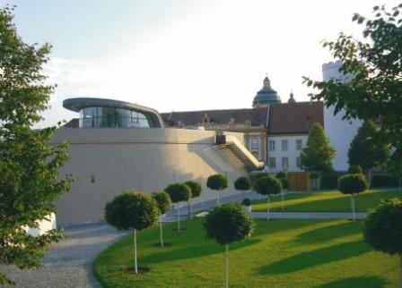 Modernes Gebäude mit geschwungener Architektur neben einem traditionellen Bauwerk, umgeben von einer Rasenfläche mit kleinen, runden Bäumen und einem gepflasterten Weg.