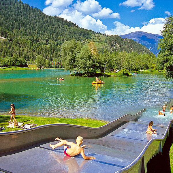 Kinder benutzen eine Wasserrutsche, die in einen klaren See führt, der von Bäumen und Bergen unter einem blauen Himmel mit Wolken umgeben ist.