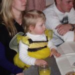 Ein kleines Kind in einem gelb-schwarzen Kostüm sitzt mit zwei Erwachsenen an einem Tisch. Auf dem Tisch stehen ein Glas mit Saft und ein Untersetzer mit der Aufschrift "Zipfer".