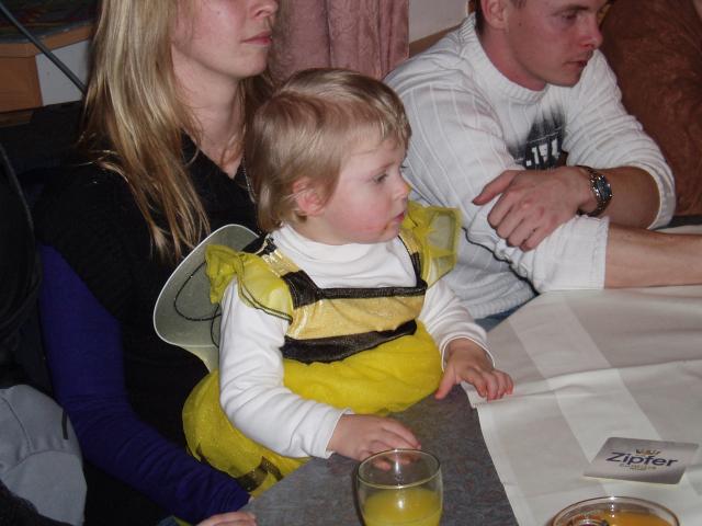 Ein kleines Kind in einem gelb-schwarzen Kostüm sitzt mit zwei Erwachsenen an einem Tisch. Auf dem Tisch stehen ein Glas mit Saft und ein Untersetzer mit der Aufschrift "Zipfer".