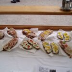 Offene Sandwiches mit verschiedenen Belägen, darunter geschnittene Zwiebeln, cremige Aufstriche, Kräuter und geschnittene Paprika, die auf weißen Servietten auf einer Holztheke angerichtet sind.