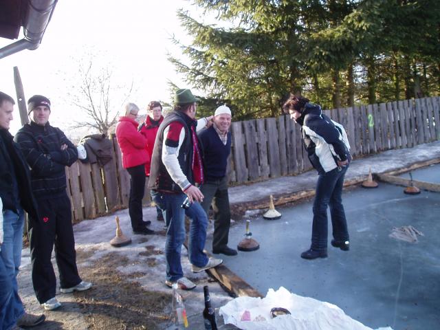 Eine Gruppe von Menschen steht an einem Holzzaun und spielt ein Spiel auf einer vereisten Fläche im Freien, mit Bäumen im Hintergrund. Einige Curlingsteine und Flaschen liegen auf dem Boden.