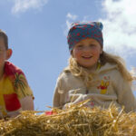 Zwei kleine Kinder, ein Junge mit Brille und ein Mädchen mit Kopftuch, lehnen lächelnd über einem Strohhaufen im Freien unter einem blauen Himmel mit Wolken.