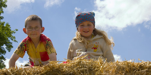 Zwei kleine Kinder, ein Junge mit Brille und ein Mädchen mit Kopftuch, lehnen lächelnd über einem Strohhaufen im Freien unter einem blauen Himmel mit Wolken.