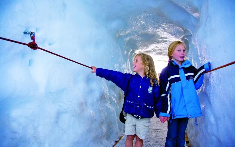 Zwei Kinder in blauen Jacken gehen durch einen Eistunnel, halten sich an den Händen und berühren ein rotes Seil an der eisigen Wand.