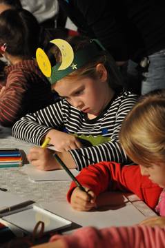 Ein Kind, das eine Papierkrone mit Mond und Sternen trägt, malt mit Buntstiften an einem Tisch zusammen mit anderen Kindern.