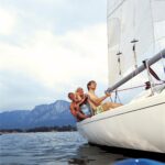 Drei Personen sitzen am Rand eines Segelboots auf einem See, im Hintergrund sind Berge und ein bewölkter Himmel zu sehen.