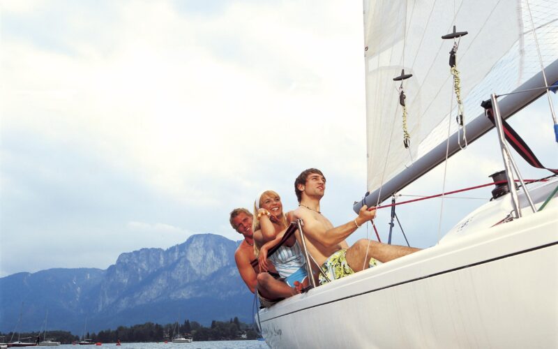 Drei Personen sitzen am Rand eines Segelboots auf einem See, im Hintergrund sind Berge und ein bewölkter Himmel zu sehen.