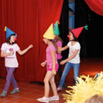 Drei Kinder mit bunten Zipfelmützen und roten Clownsnasen treten auf der Bühne vor einem roten Vorhang auf, während andere Kinder sitzen und zusehen.