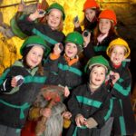 Eine Gruppe von Kindern mit Schutzhelmen und Bergbaujacken posiert in einer Höhle mit einer Zwergenstatue vor sich.