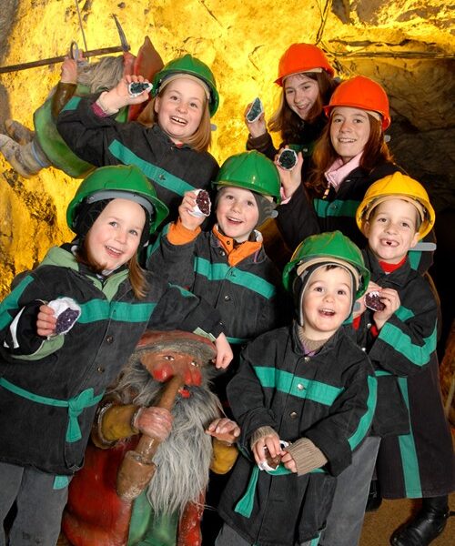 Eine Gruppe von Kindern mit Schutzhelmen und Bergbaujacken posiert in einer Höhle mit einer Zwergenstatue vor sich.