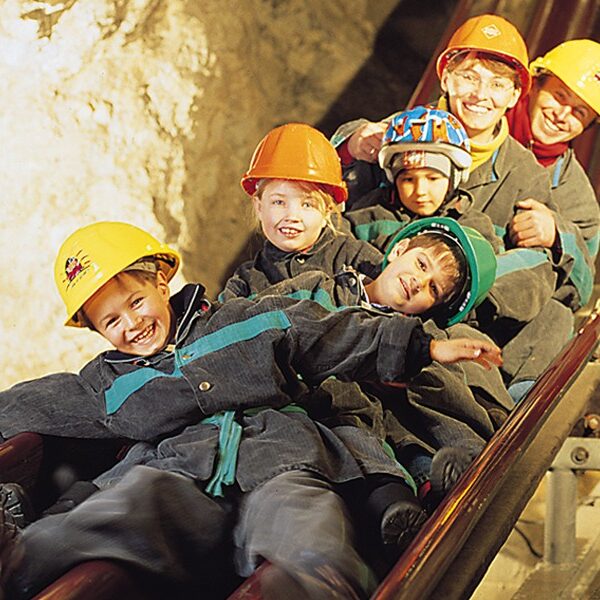 Eine Gruppe von Kindern und Erwachsenen mit Helmen und Schutzanzügen rutscht eine Holzrutsche in einer Höhle oder einem Bergwerk hinunter.
