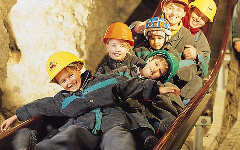 Eine Gruppe von Kindern und Erwachsenen mit Helmen und Schutzanzügen rutscht eine Holzrutsche in einer Höhle oder einem Bergwerk hinunter.