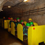 Menschen mit Schutzhelmen fahren in einem kleinen gelben Zug durch einen unterirdischen Tunnel mit felsigen Wänden und Oberlichtern.