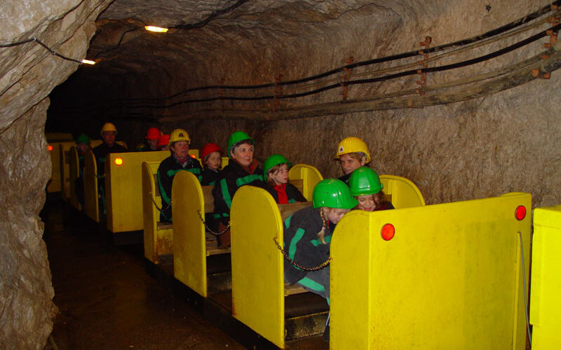Menschen mit Schutzhelmen fahren in einem kleinen gelben Zug durch einen unterirdischen Tunnel mit felsigen Wänden und Oberlichtern.