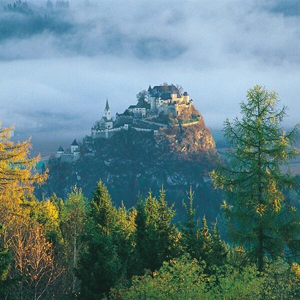 Ein Schloss thront auf einem felsigen Hügel, umgeben von Bäumen und Nebel, und im Hintergrund sind schwach die Berge zu erkennen.