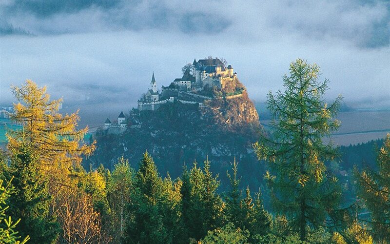 Ein Schloss thront auf einem felsigen Hügel, umgeben von Bäumen und Nebel, und im Hintergrund sind schwach die Berge zu erkennen.