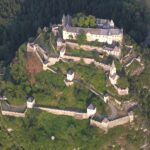 Luftaufnahme einer großen mittelalterlichen Burganlage mit mehreren Mauern, Türmen und Gebäuden, umgeben von dichtem Wald.