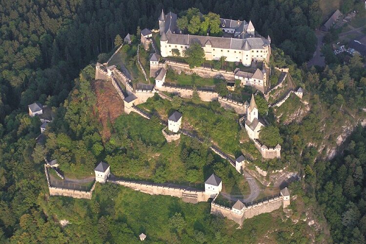Luftaufnahme einer großen mittelalterlichen Burganlage mit mehreren Mauern, Türmen und Gebäuden, umgeben von dichtem Wald.