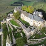 Luftaufnahme einer großen mittelalterlichen Burganlage, umgeben von Steinmauern, Türmen und Bäumen, gelegen auf einem Hügel inmitten von Feldern und Wäldern.