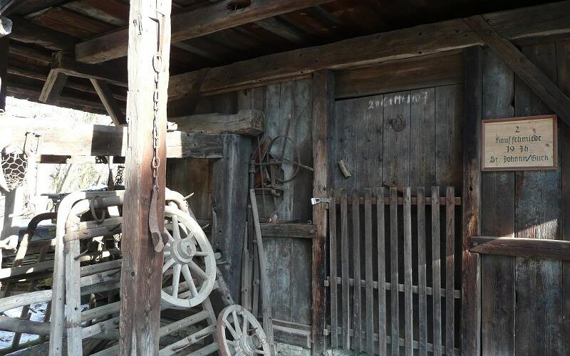 Holzschuppen mit rustikalen Werkzeugen und Geräten, einer vergitterten Holztür und einem Schild mit der Aufschrift "2. Samalmador 19 St. Johann/Verns". Das Sonnenlicht wirft Schatten auf das verwitterte Gebäude.