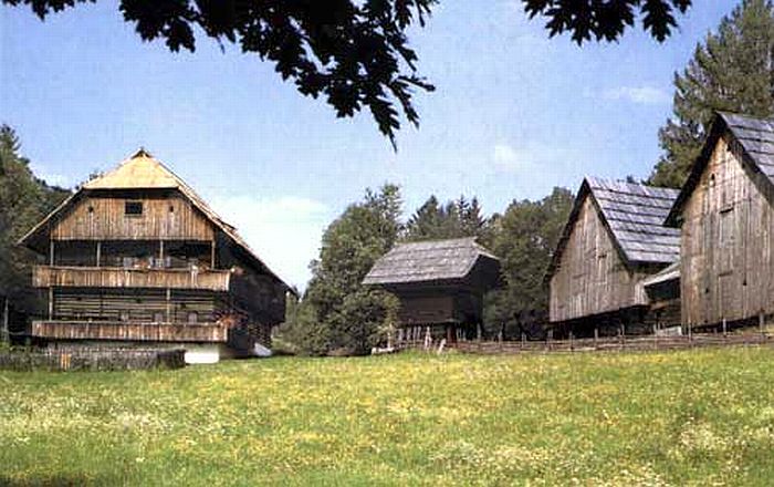 Drei traditionelle hölzerne Bauernhäuser stehen auf einer von Bäumen umgebenen Wiese unter einem klaren blauen Himmel.