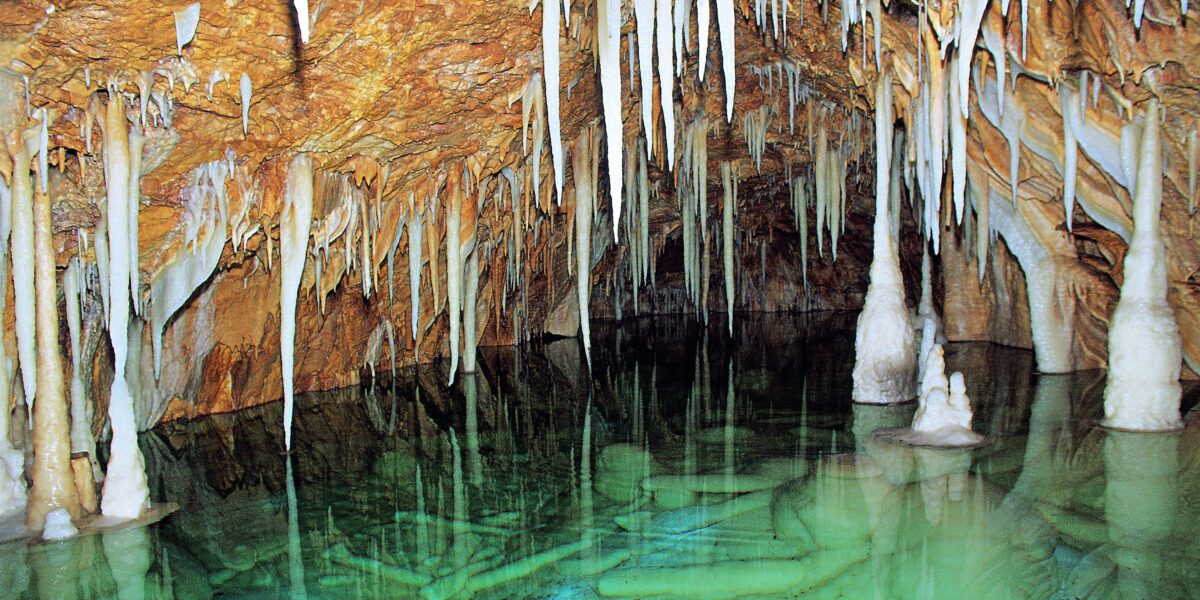 Unterirdische Höhle mit hängenden Stalaktiten, stehenden Stalagmiten und klarem grünen Wasser, in dem sich die Formationen spiegeln.