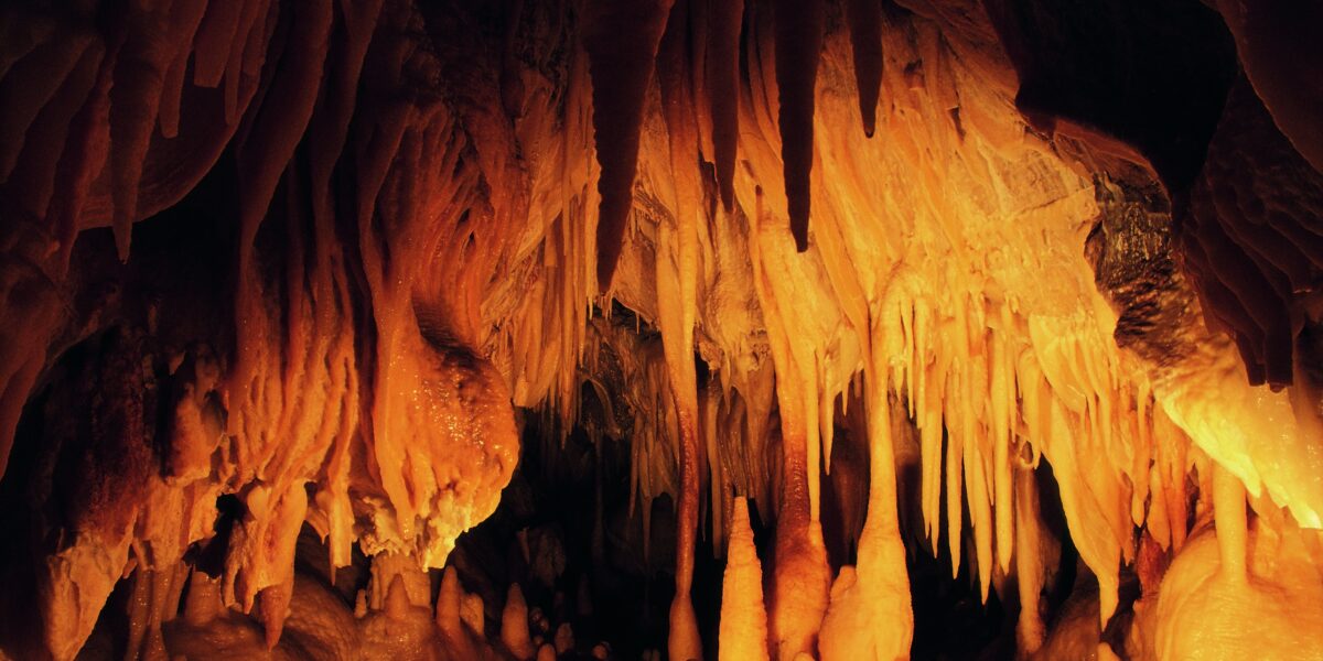 Ein Höhleninneres mit zahlreichen orangefarbenen Stalaktiten, die von der Decke hängen, und Stalagmiten, die aus dem Boden ragen, beleuchtet von warmem Licht.