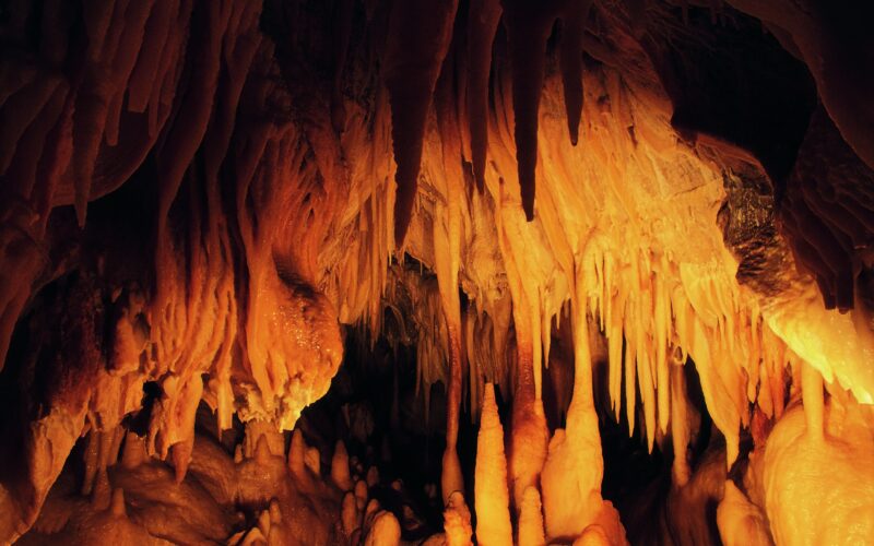Ein Höhleninneres mit zahlreichen orangefarbenen Stalaktiten, die von der Decke hängen, und Stalagmiten, die aus dem Boden ragen, beleuchtet von warmem Licht.