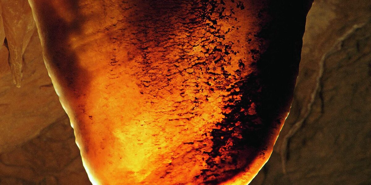 Eine Nahaufnahme einer durchscheinenden, orange-braunen Höhlenformation, möglicherweise ein Stalaktit oder Vorhang, beleuchtet vor einem dunklen Hintergrund.